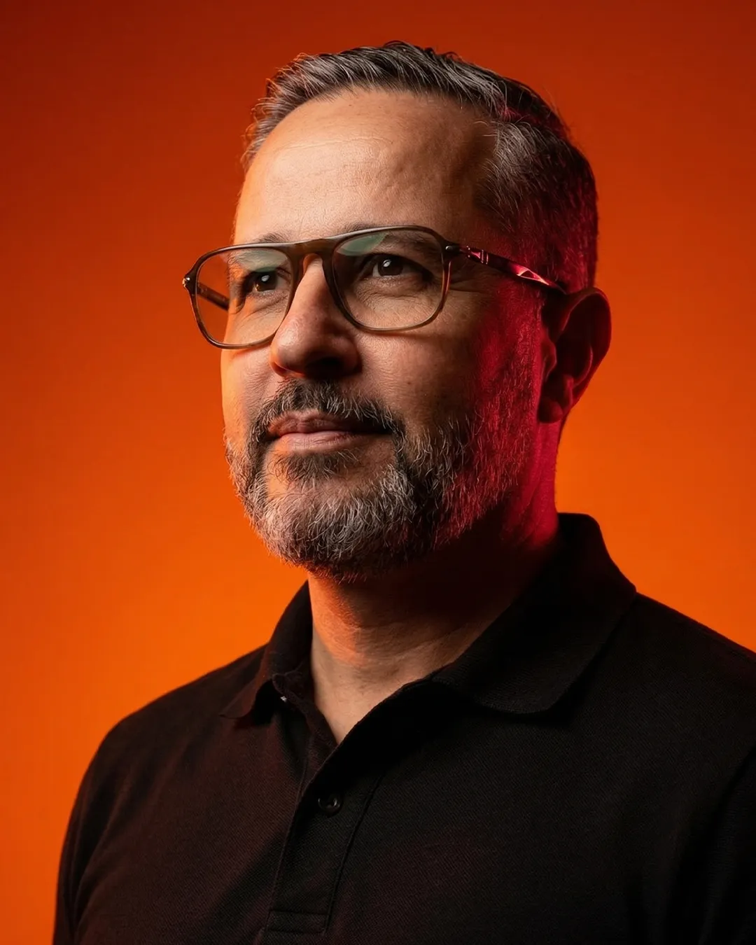 Bearded man with glasses wearing a black shirt against a warm orange background, looking thoughtful.