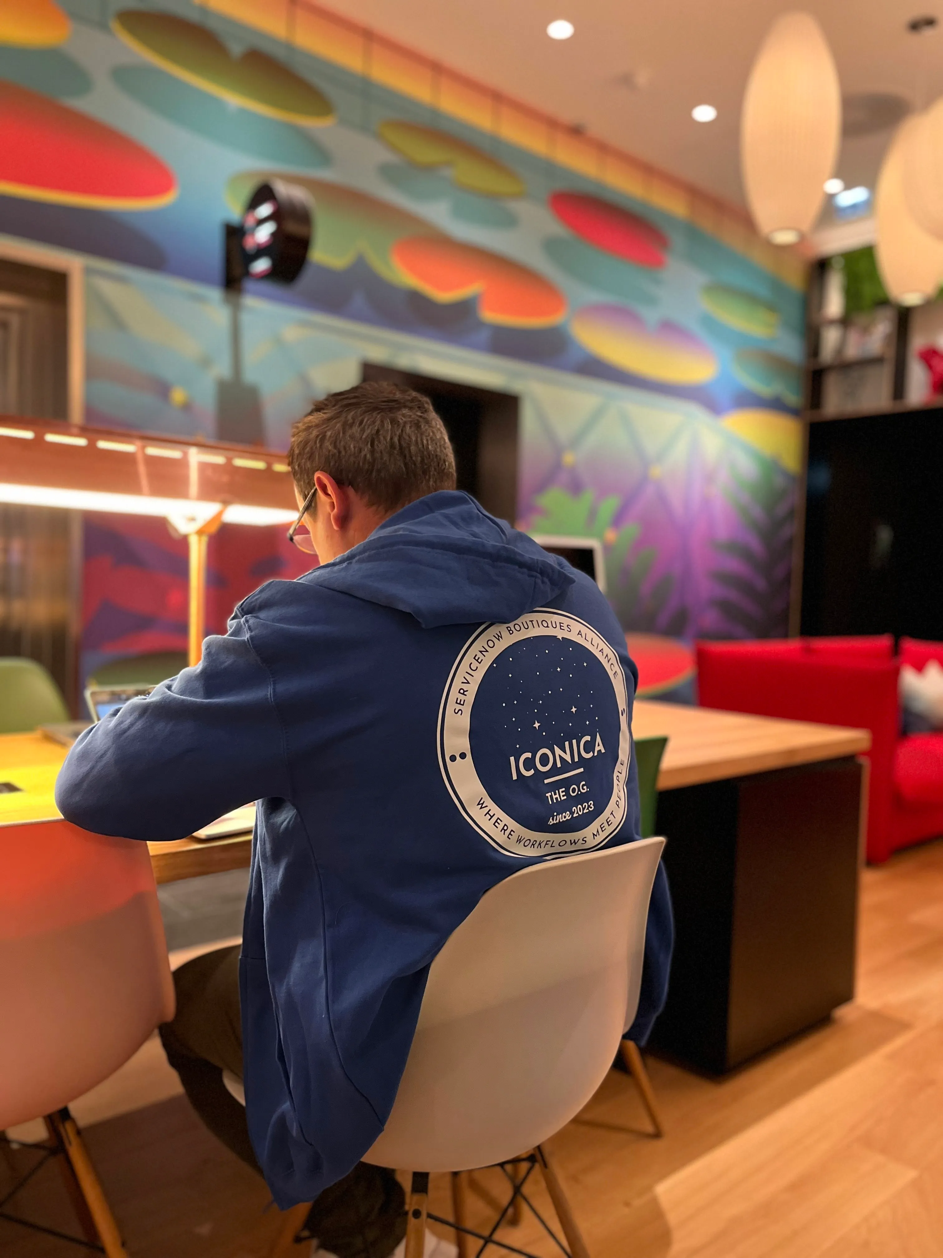 Person wearing blue hoodie with ICONICA logo working on a laptop at a colorful modern workspace.