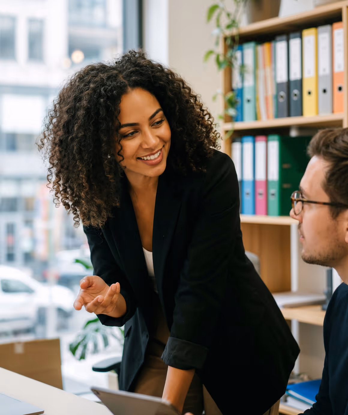 Smiling woman in black blazer engaging in conversation with seated man in office.