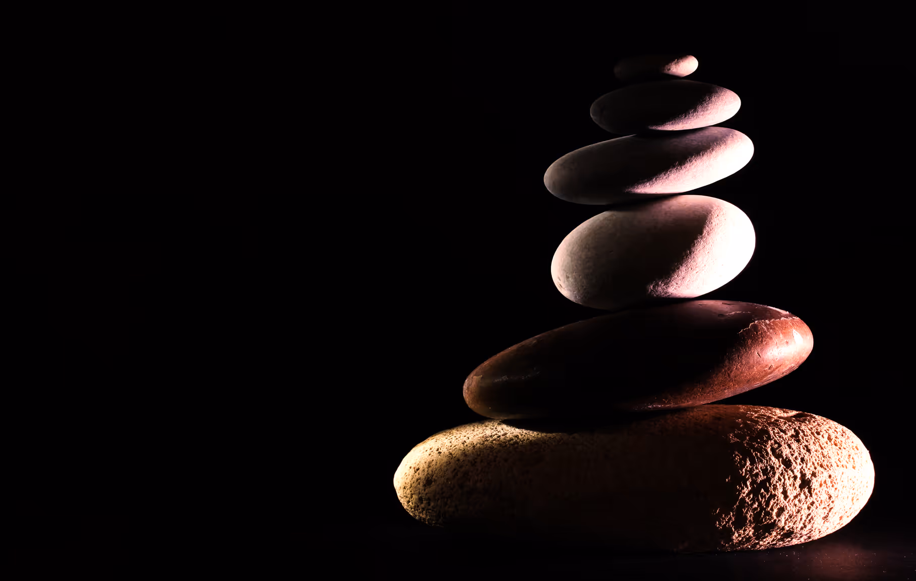 Stack of seven smooth stones balanced against a black background with soft lighting highlighting their textures.