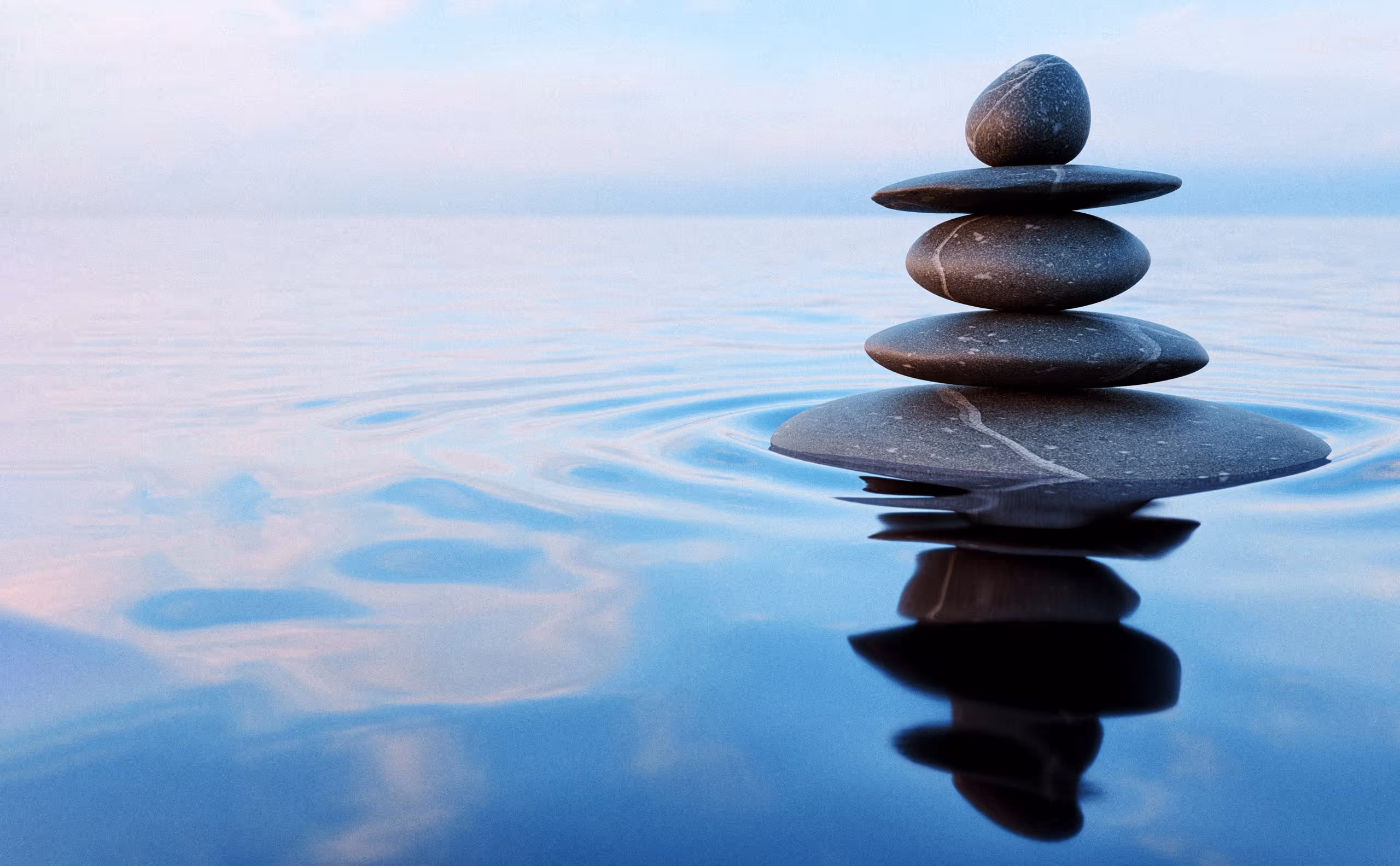 Four smooth stones balanced in a stack on calm water with their reflection visible and a soft sky background.