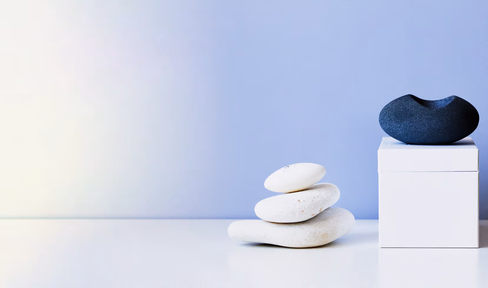 Three smooth white stones stacked beside a dark stone on a white cube against a soft blue background.