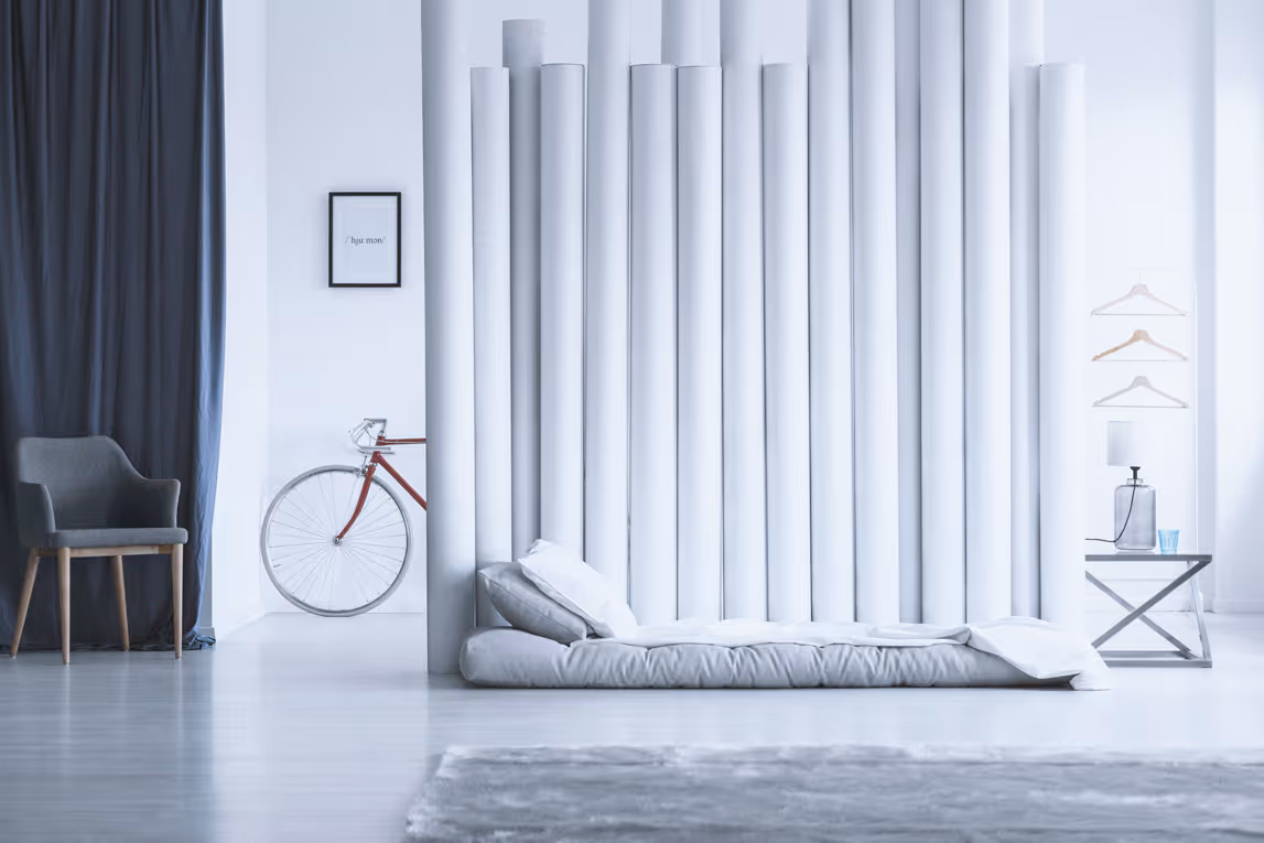 Minimalist white bedroom with low platform bed, gray chair, bicycle, and hanging clothes hangers.