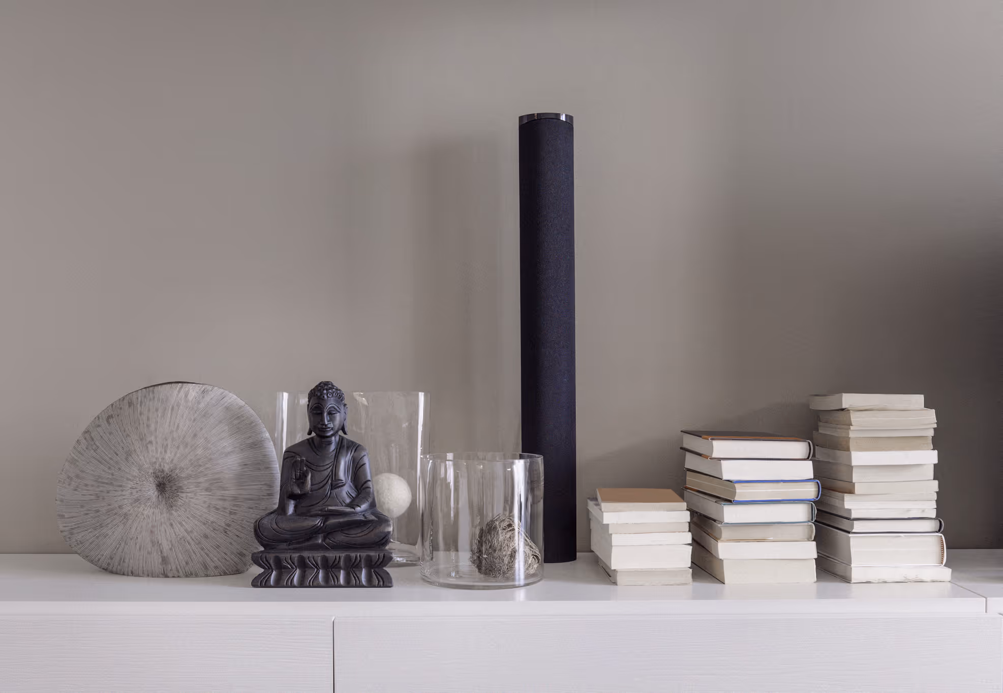 Modern white shelf with a black Buddha statue, decorative glass vases, black cylindrical speaker, and stacks of books against a gray wall.