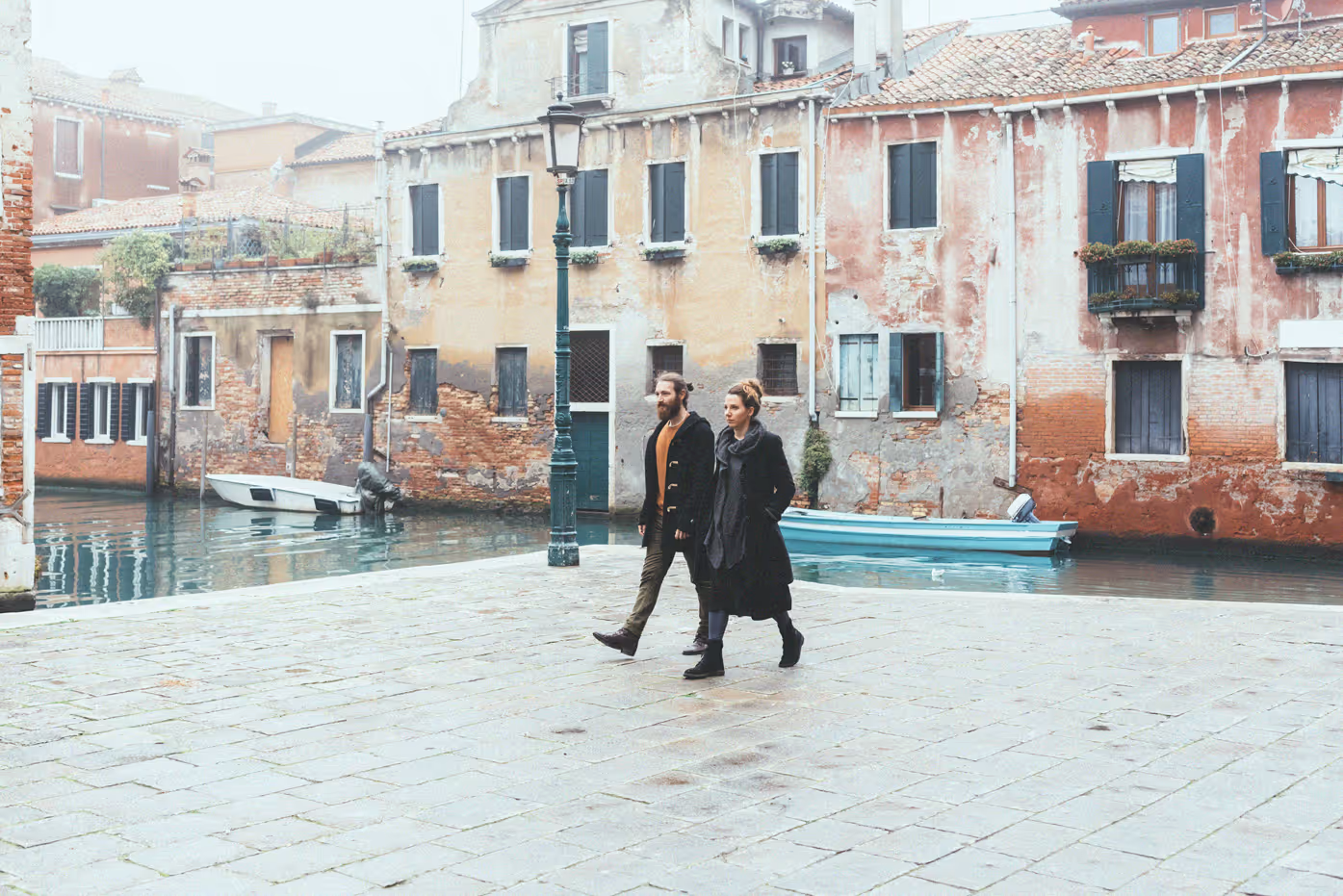 A man and woman dressed in dark coats walking on a stone pavement beside a canal with old, weathered buildings in the background.