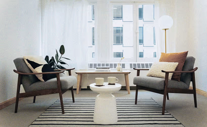Minimalist living room with two gray armchairs with pillows, a wooden coffee table, a small white side table with two mugs, a striped rug, and a floor lamp by a window.