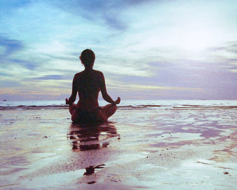 Person in a meditative pose sitting cross-legged on a wet beach at sunrise or sunset.