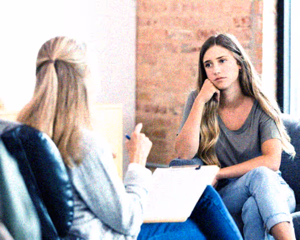 Young woman sitting on a couch looking thoughtfully at a therapist holding a clipboard.