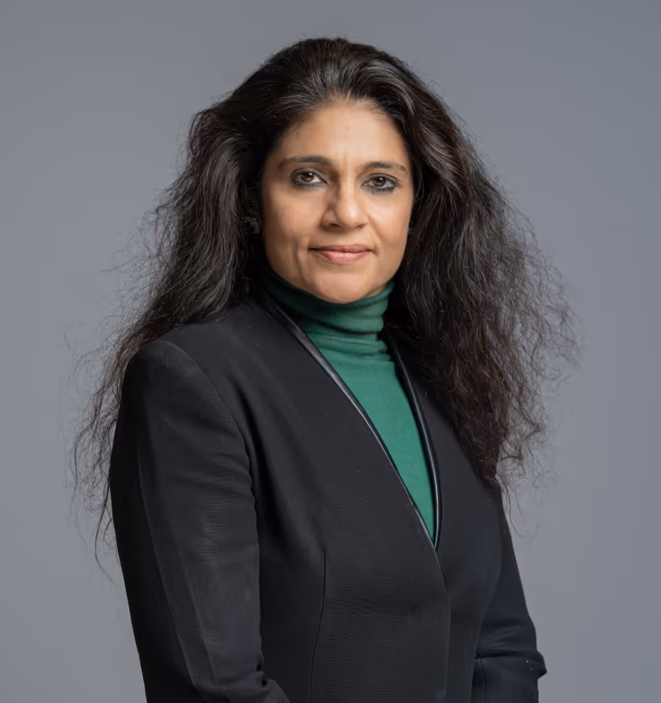 Woman with long dark hair wearing a black blazer over a green turtleneck standing against a gray background.