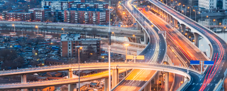 Nighttime city highway interchange with light trails from moving vehicles and surrounding buildings.