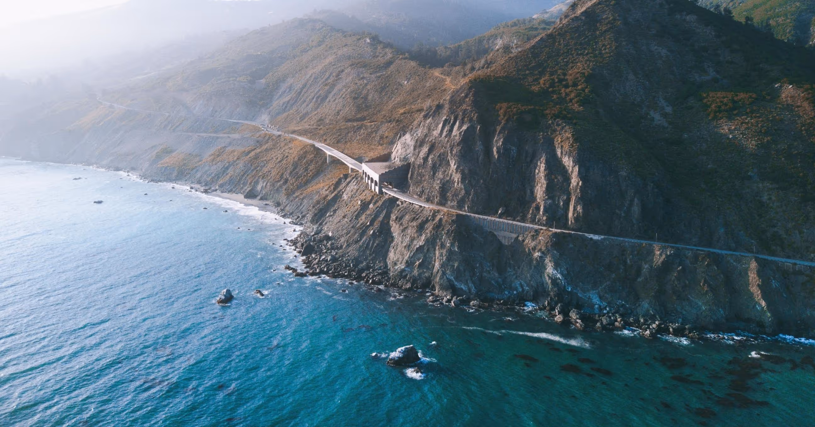 Coastal highway winding along rugged cliffs with blue ocean water below and misty hills in the background.