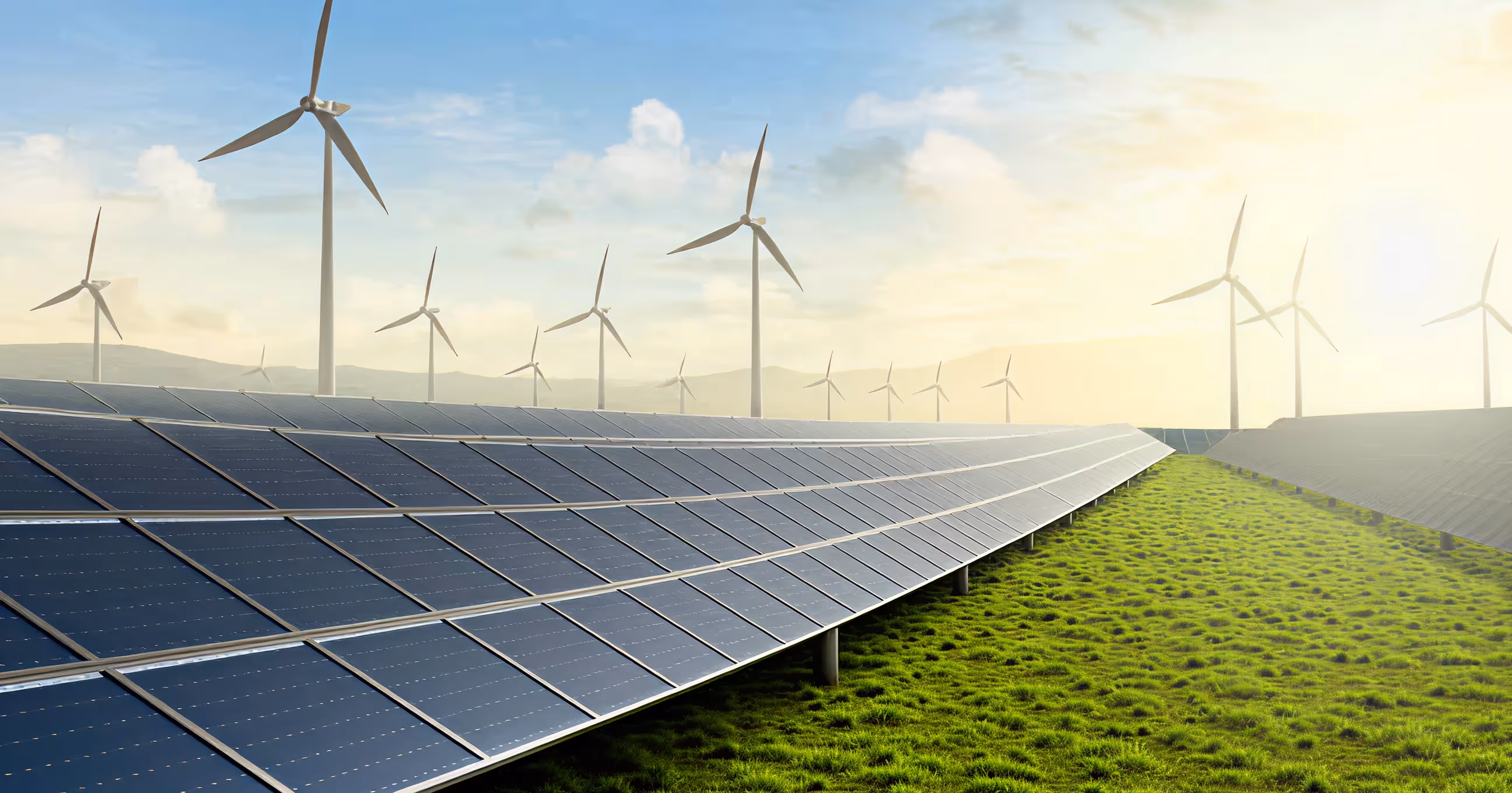 Solar panels on green grass field with wind turbines under a partly cloudy sky at sunrise or sunset.