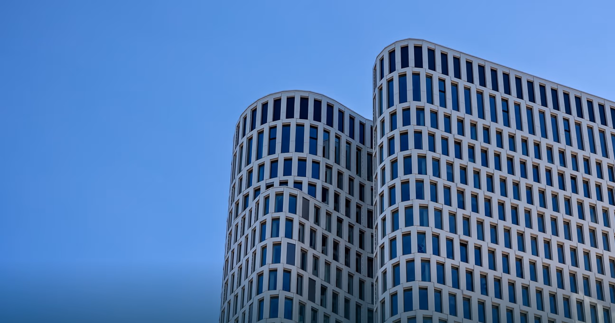 Modern high-rise building with curved corners and a grid of large blue-tinted windows under a clear blue sky.