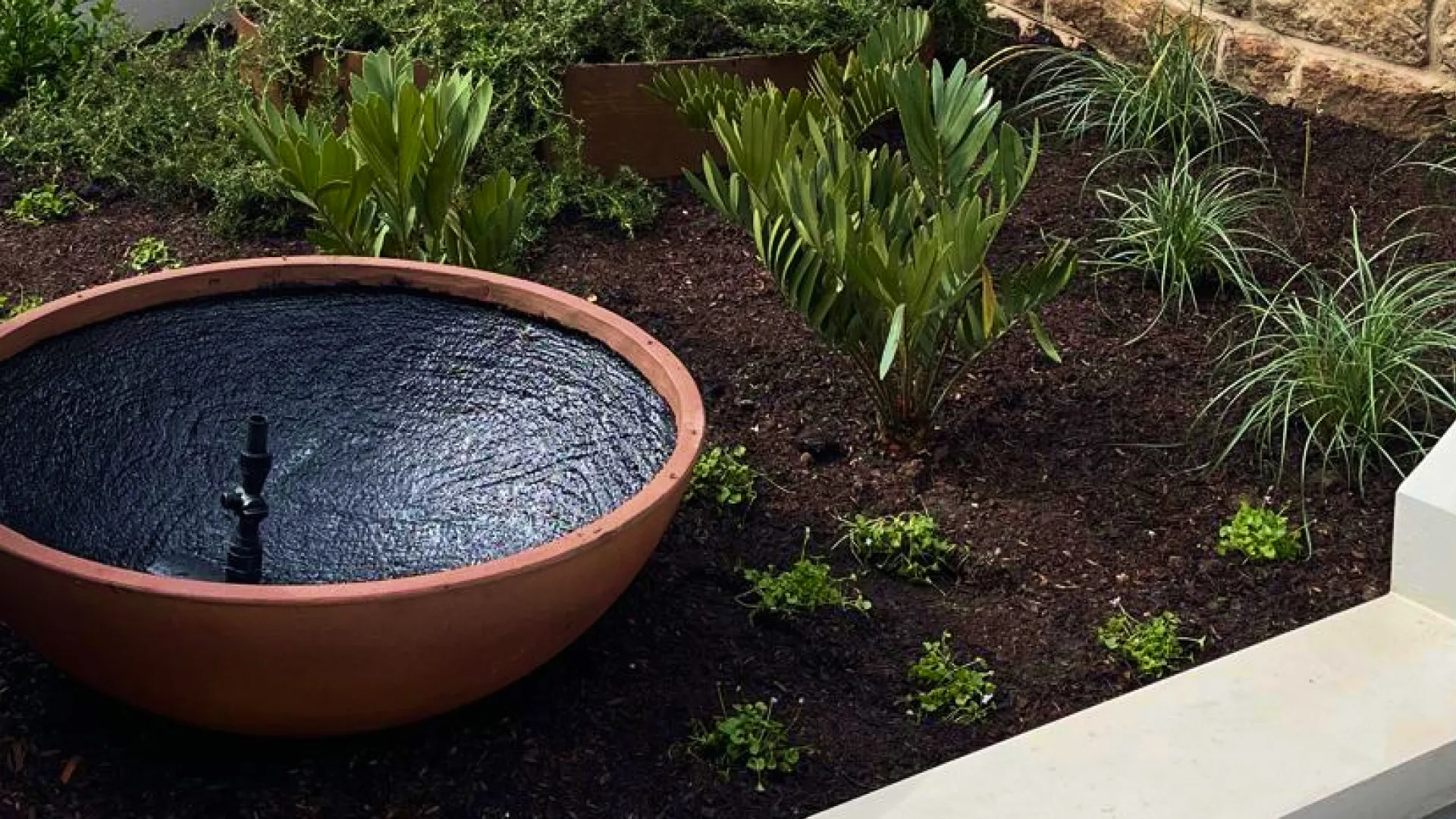 Terracotta garden fountain with water feature surrounded by green plants and dark soil.