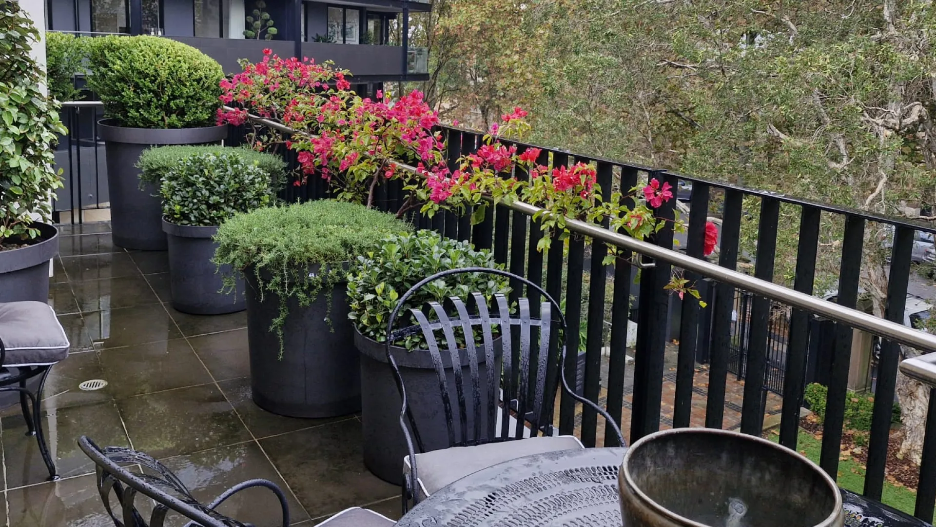 Balcony with large planters with green plants, and pink flowers on the railing.