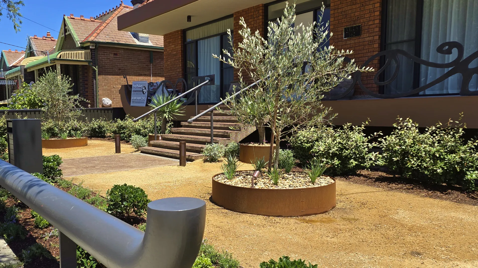 Residential area with dry garden beds, small shrubs and olive trees.