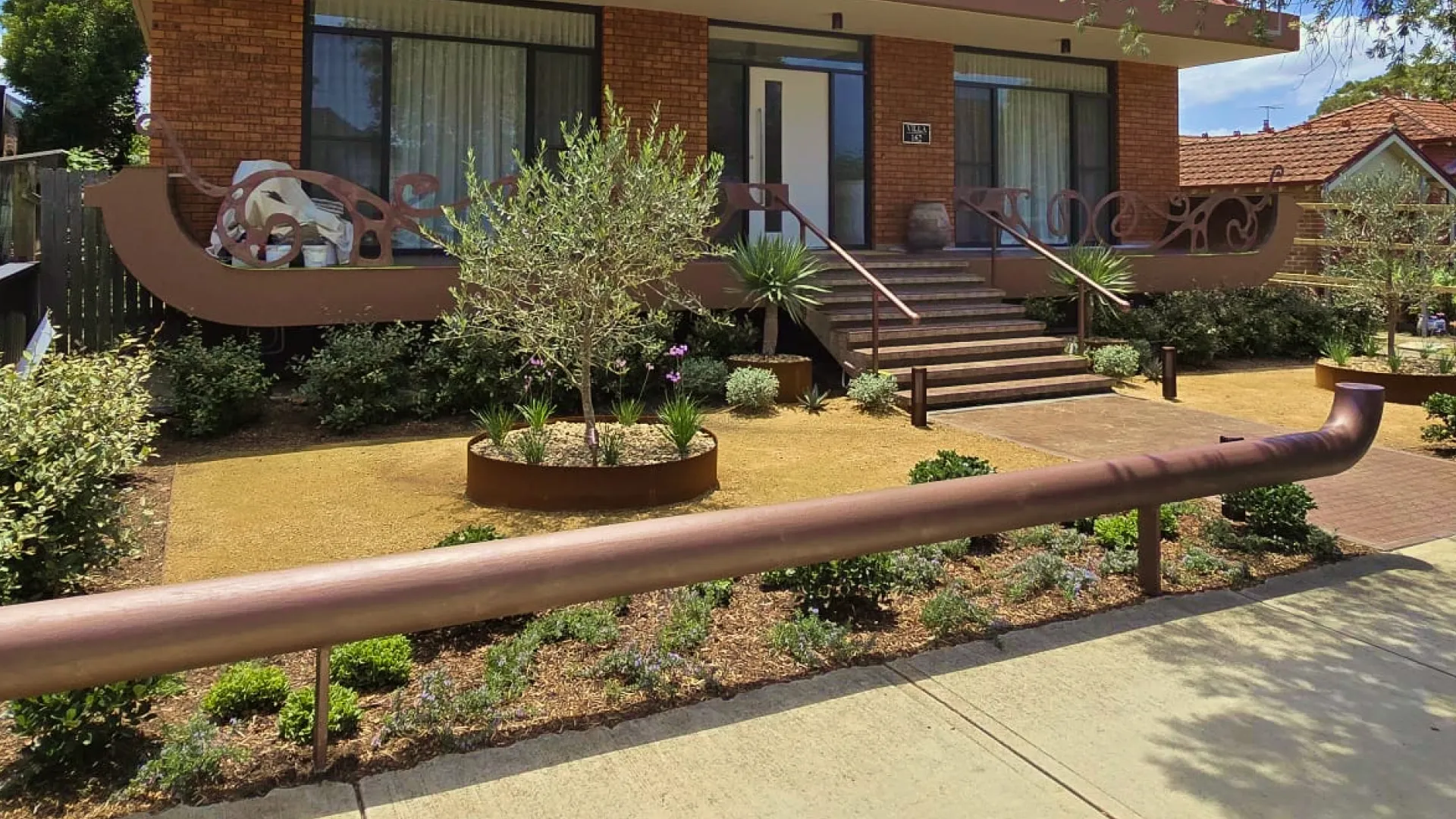 Residential area with dry garden beds, small shrubs, olive trees, brown railing, and a concrete sidewalk.