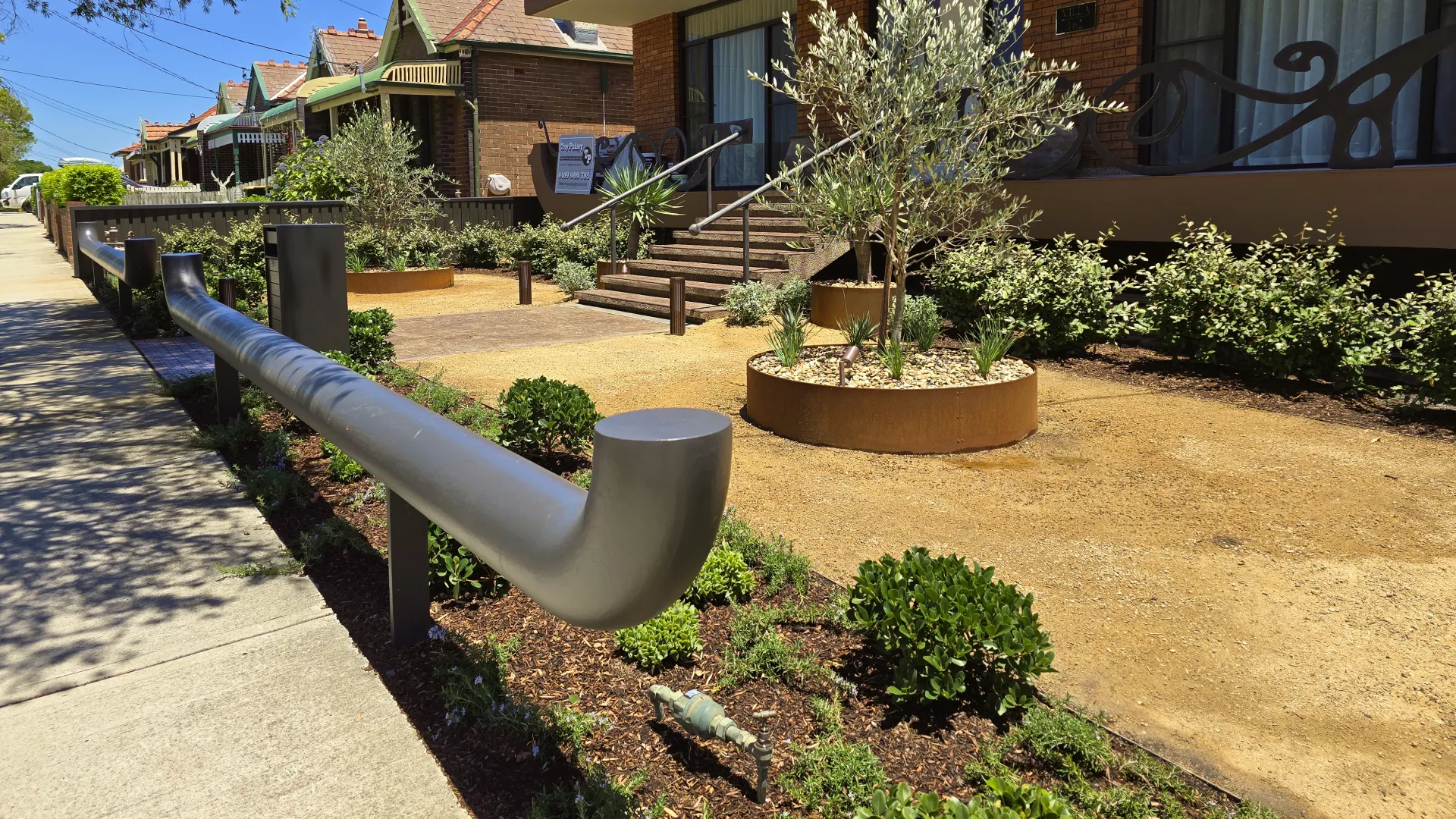 Residential area with dry garden beds, small shrubs, olive trees, brown railing, and a concrete sidewalk.