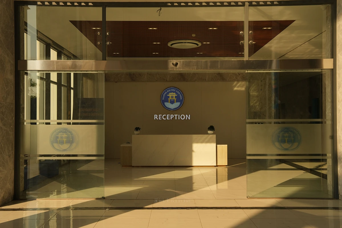 Üres, üvegfalú recepciós tér napfényes árnyékokkal, középen pulttal és a falon „RECEPTION” felirattal.