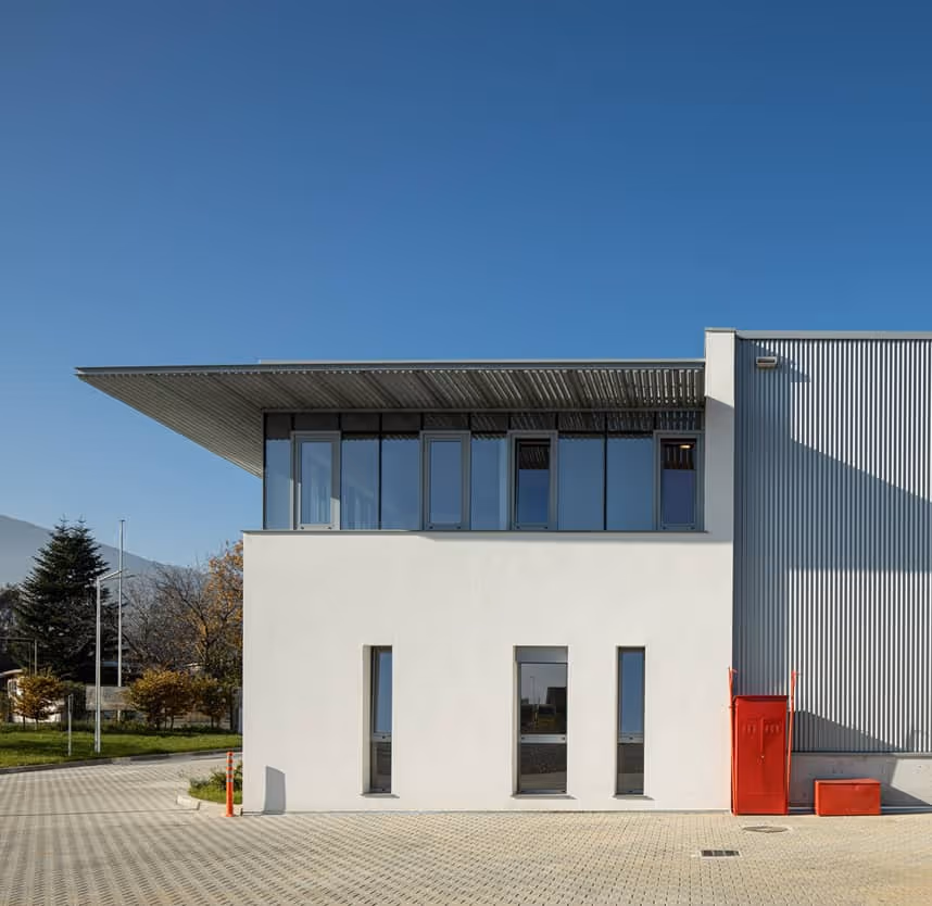 Modern white and gray industrial building with large windows and red fire safety equipment outdoors under clear blue sky.