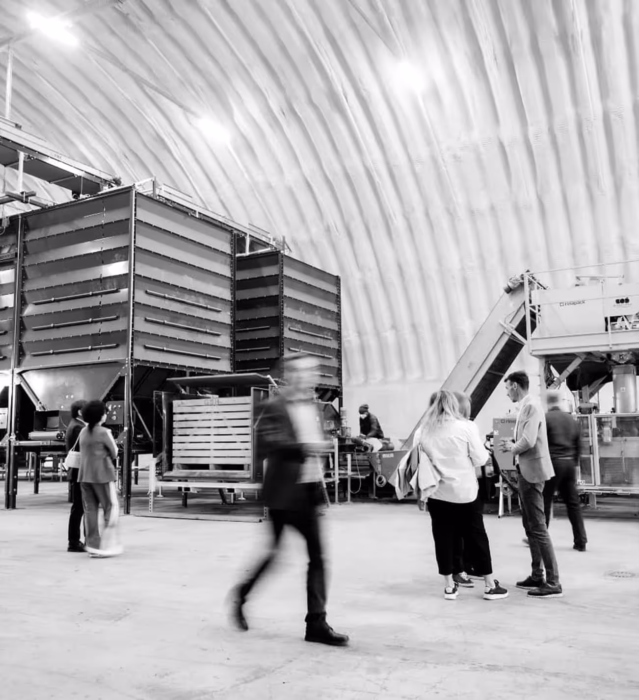 People standing and walking inside a large industrial facility with machinery and equipment under a high ceiling.