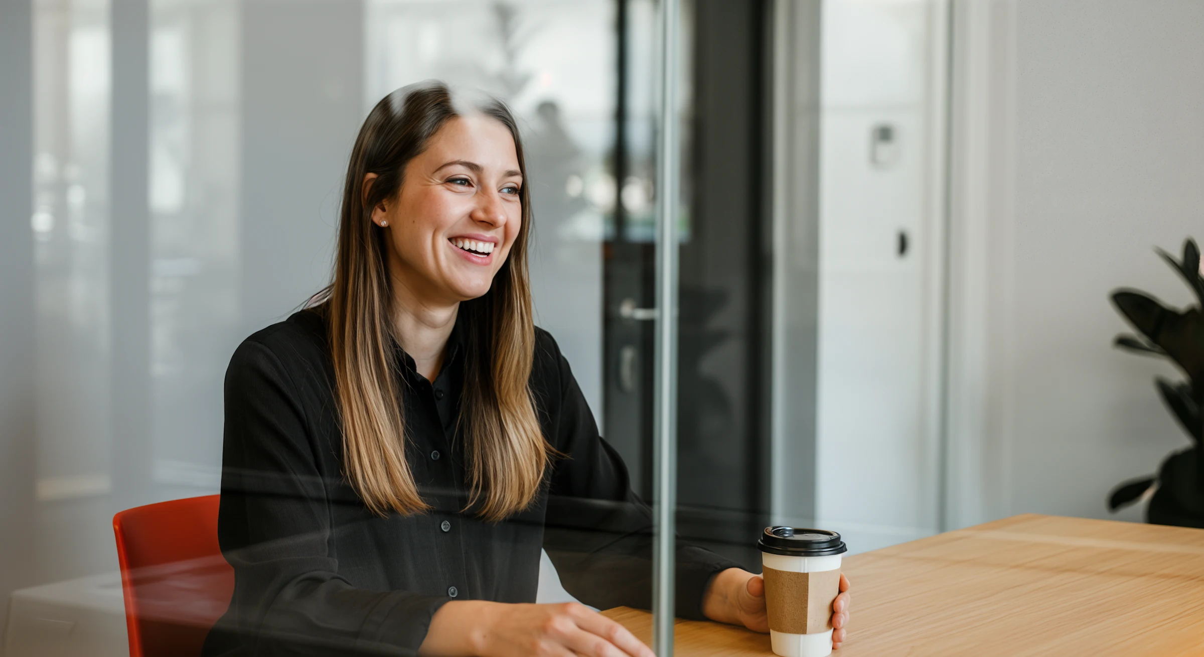Smiling woman in a black shirt sitting at a table holding a takeaway coffee cup.