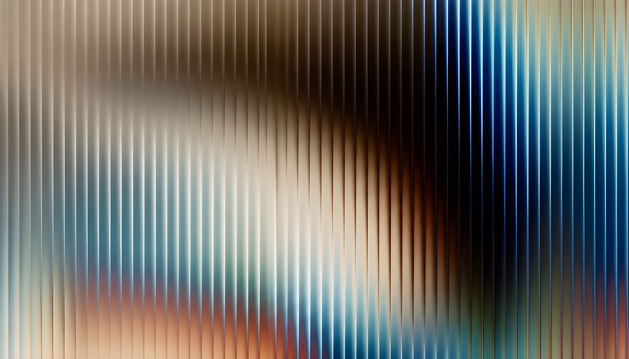 Abstract image of vertical ribbed glass or reeded glass panel with a soft gradient of colors — creamy white and beige on the left transitioning to deep navy blue on the right, with warm amber and rust tones at the center and lower portions, creating a blurred, luminous effect through the textured surface.
