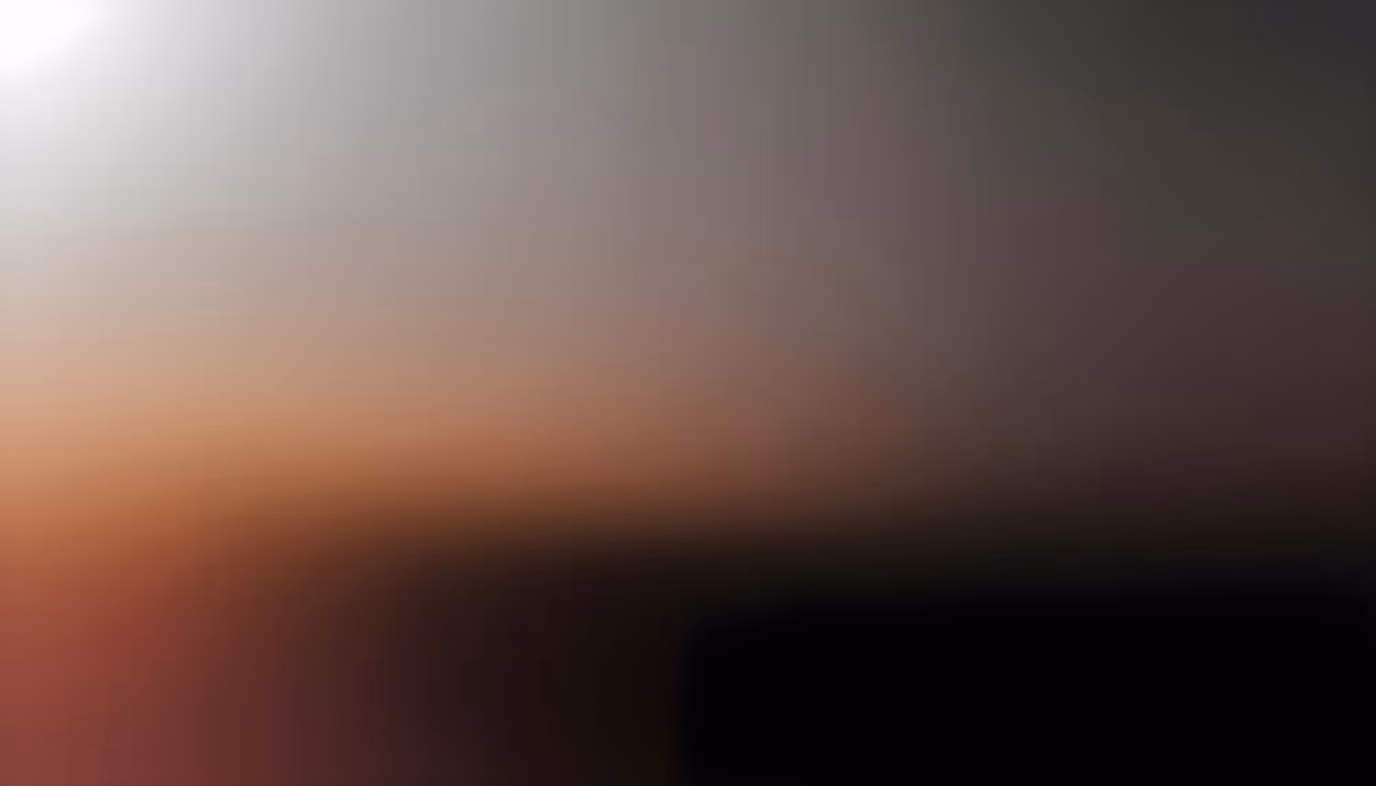Heavily blurred abstract image with warm orange and brown tones in the lower left, fading to dark brown and black on the right, with a bright white light source in the upper left corner against a gray background.