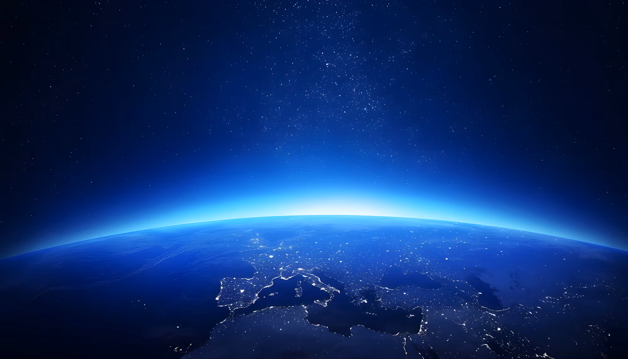 Earth viewed from space at night, showing Europe and the Mediterranean region illuminated by city lights against a deep blue ocean, with the glowing blue arc of the atmosphere and a star-filled sky above.