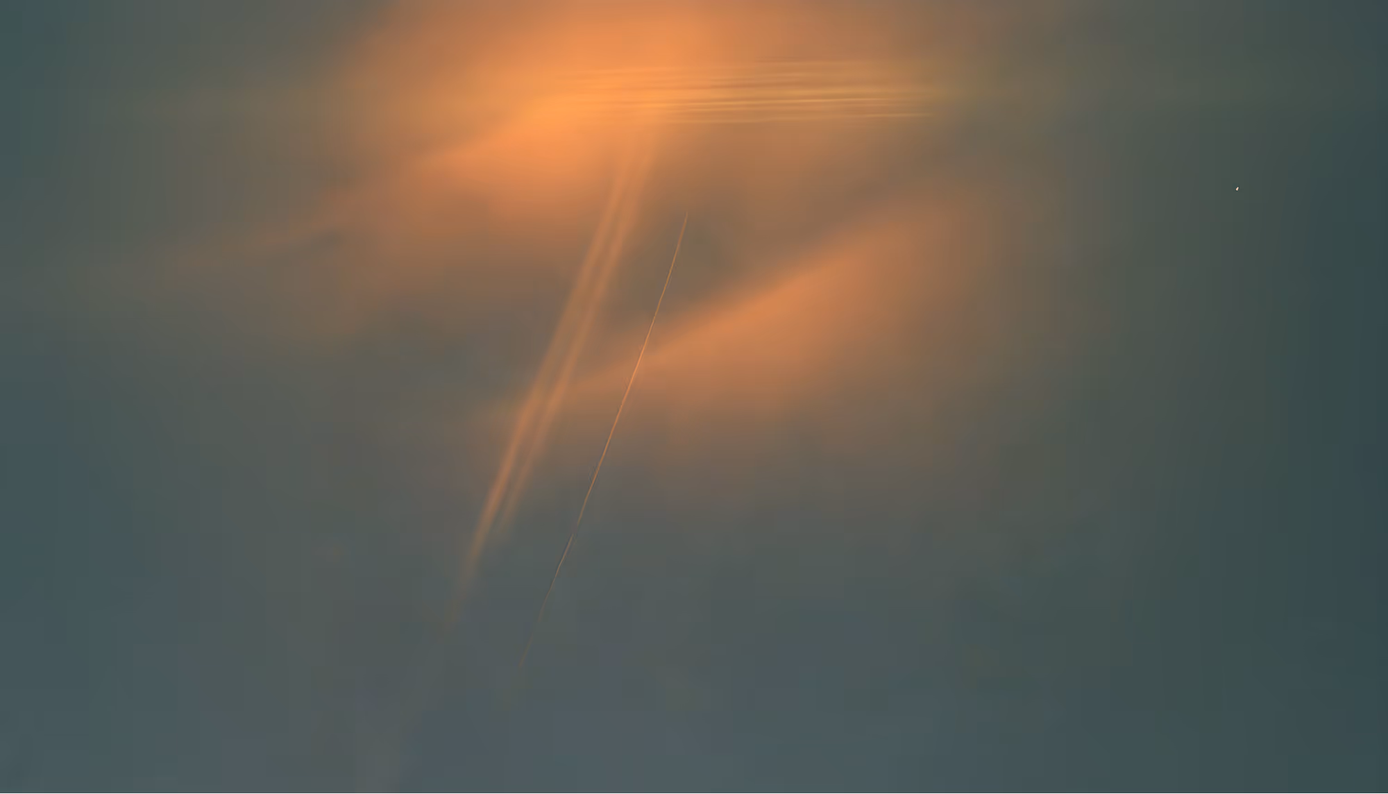 A plane soars through a dusky sky, leaving a thin contrail. Sunlight casts a warm orange hue on wispy clouds, creating a tranquil and ethereal atmosphere.