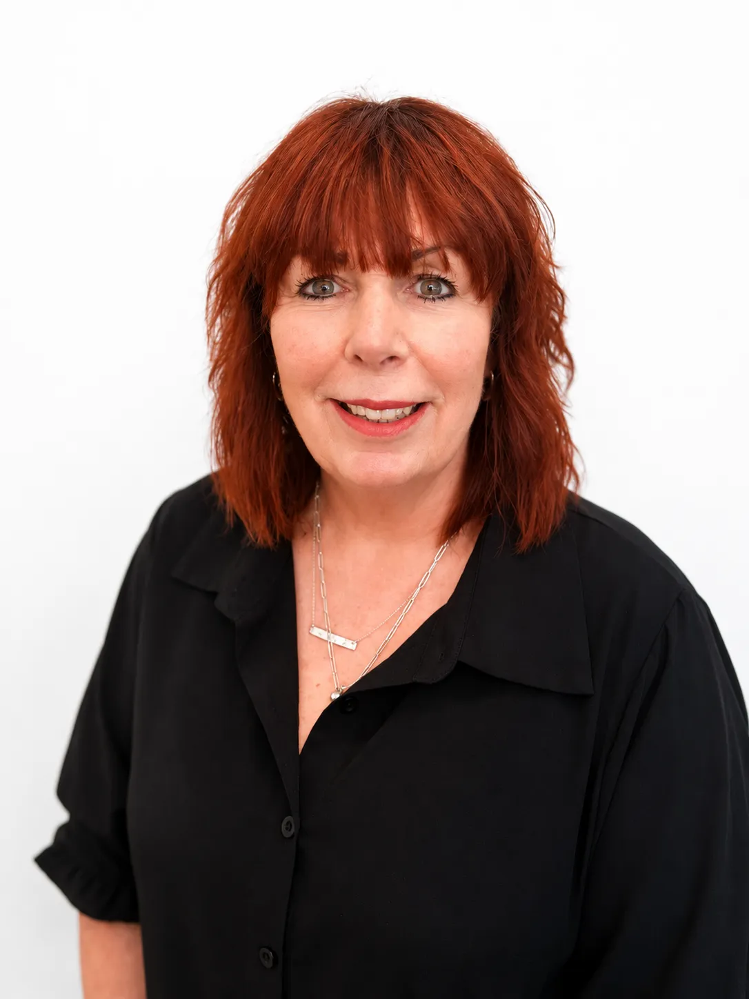 Smiling woman with shoulder-length red hair wearing a black shirt and silver necklaces on white background.