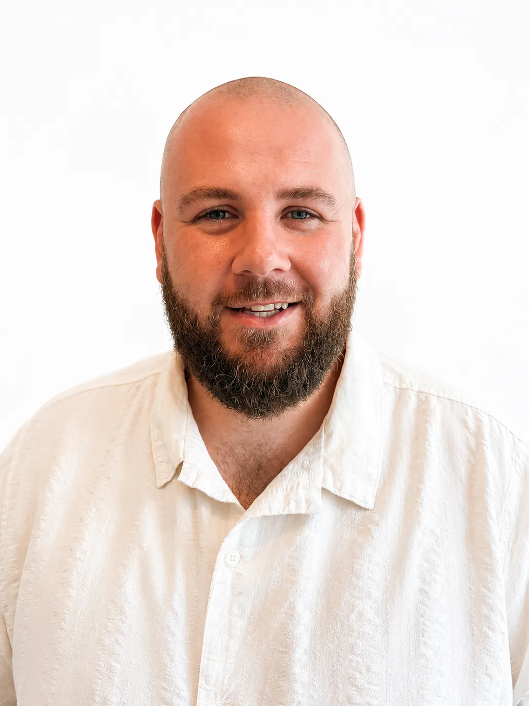 Smiling bald man with a full beard wearing a textured white button-up shirt against white background.