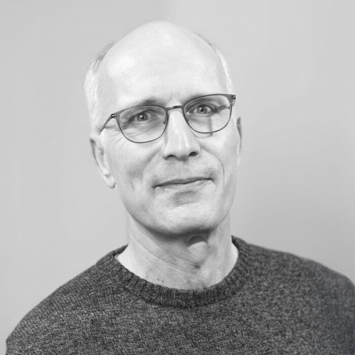 Portrait of an older man with glasses wearing a dark sweater, smiling softly against a plain background.
