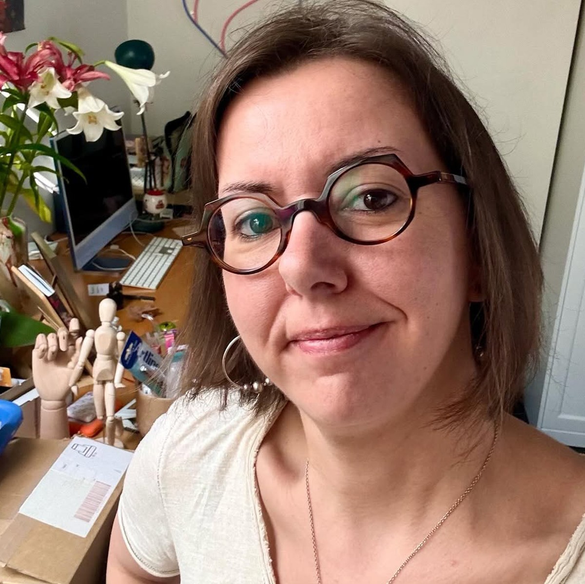 Woman with short brown hair and round glasses smiling in a cluttered room with flowers and art supplies.