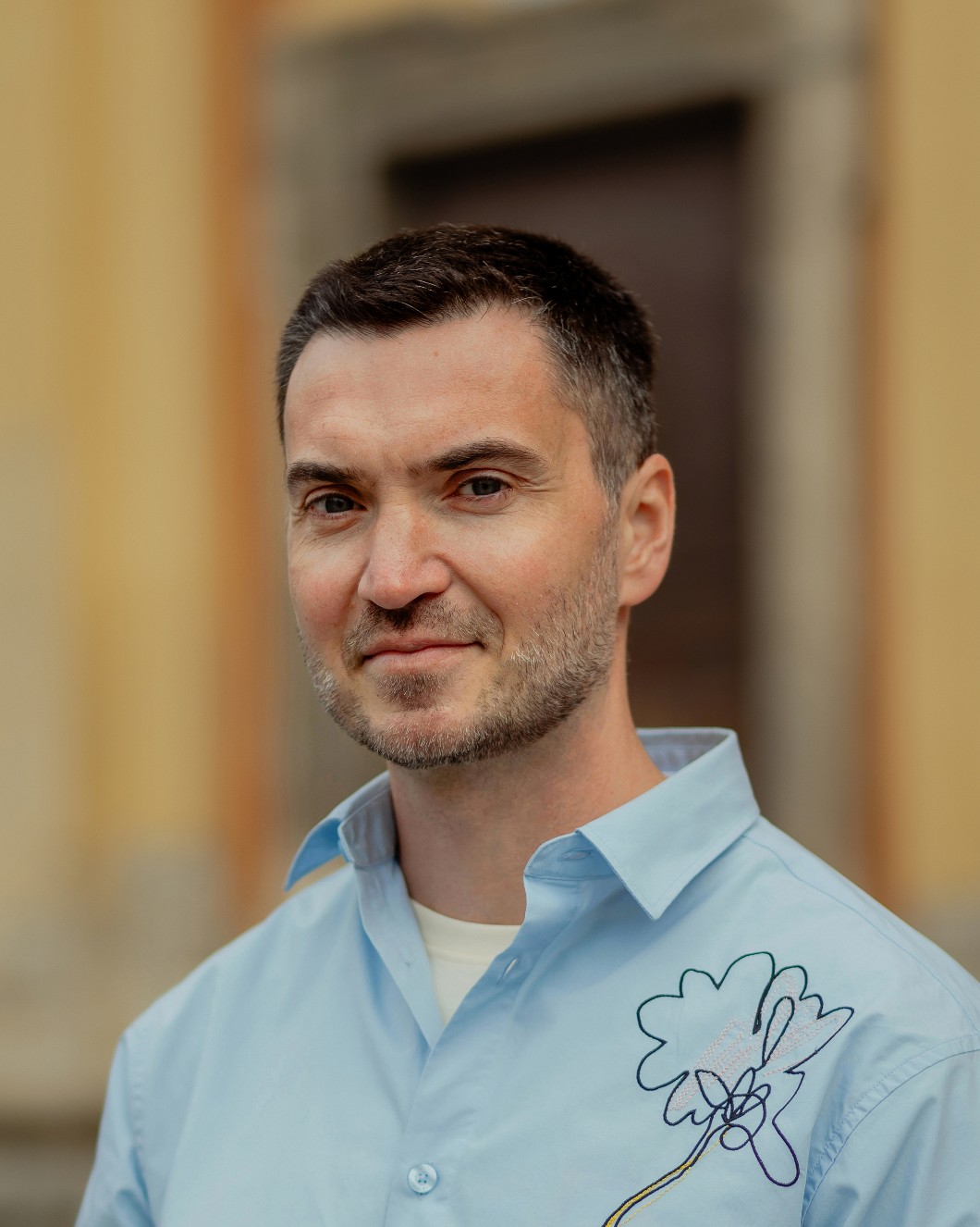 Man with short dark hair and stubble wearing a light blue shirt with a floral embroidery.