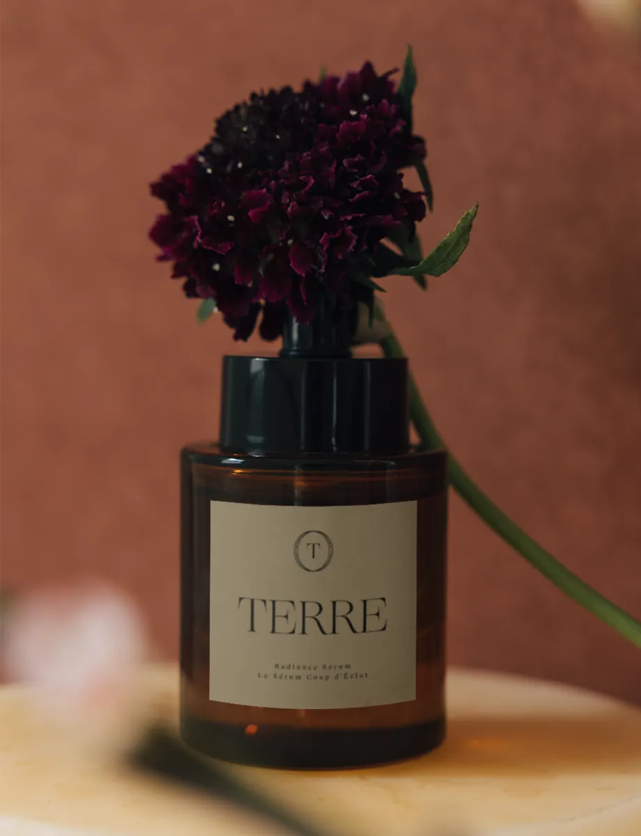 Branding-Konzept für TERRE, eine Premium-Skincare-Linie – Radiance Serum in Amber-Glasflasche mit cremefarbenem Label, Serif-Logo und T-Signet, vor warmem Terrakotta-Hintergrund.