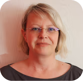 Porträt einer lächelnden Frau mit kurzen blonden Haaren, die eine Brille trägt, vor hellem Hintergrund.