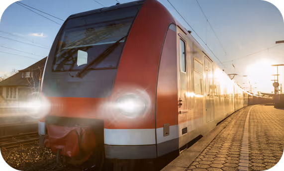 Ein roter Zug mit eingeschaltetem Scheinwerfer steht am Bahnsteig bei Sonnenuntergang.