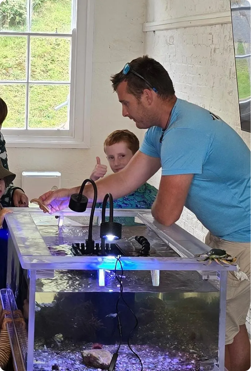 Man feeding a small animal from a clear water tank while a child gives a thumbs up in the background.