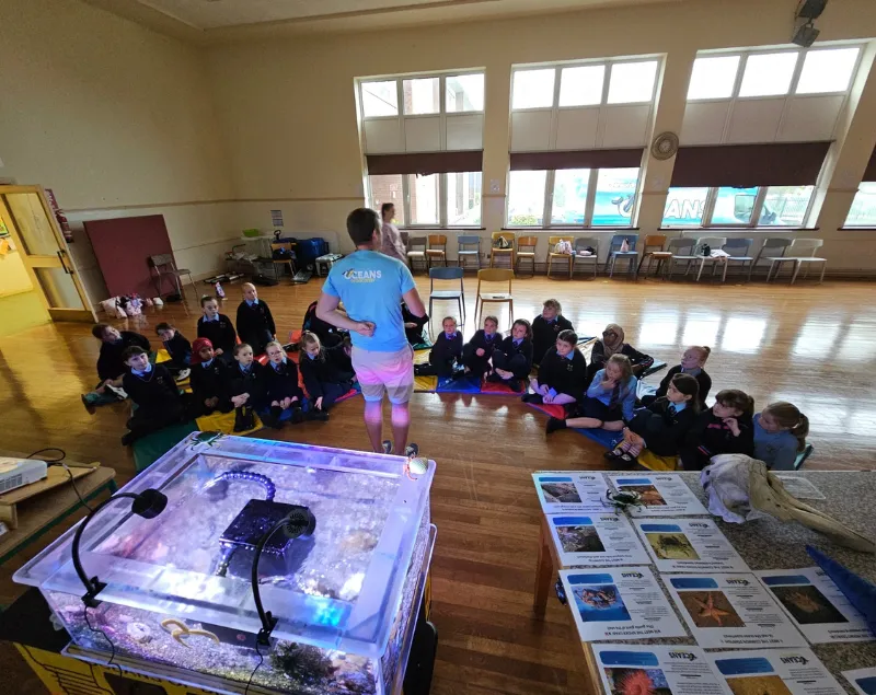 Adult instructor standing in front of seated children in a large room with aquarium tanks and educational materials on tables.