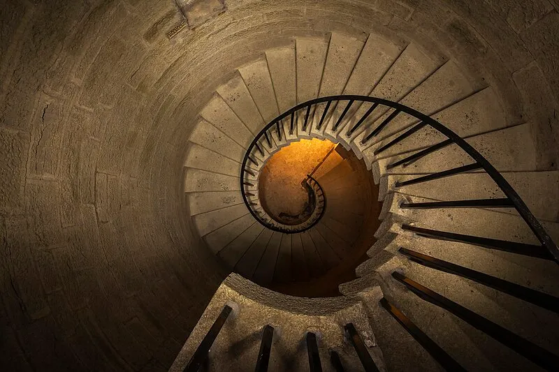 View looking down a stone spiral staircase with a metal handrail and warm lighting at the bottom.