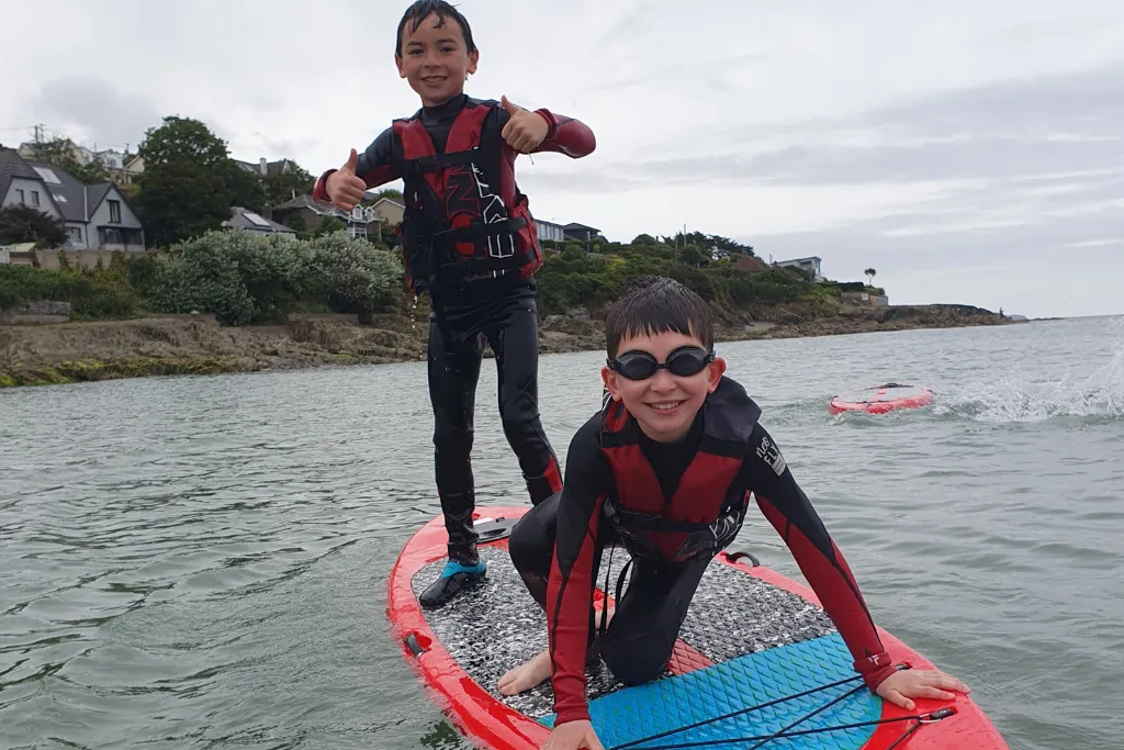 Two boys in wetsuits and life jackets on a red paddleboard in calm water near a rocky shore with houses and greenery.