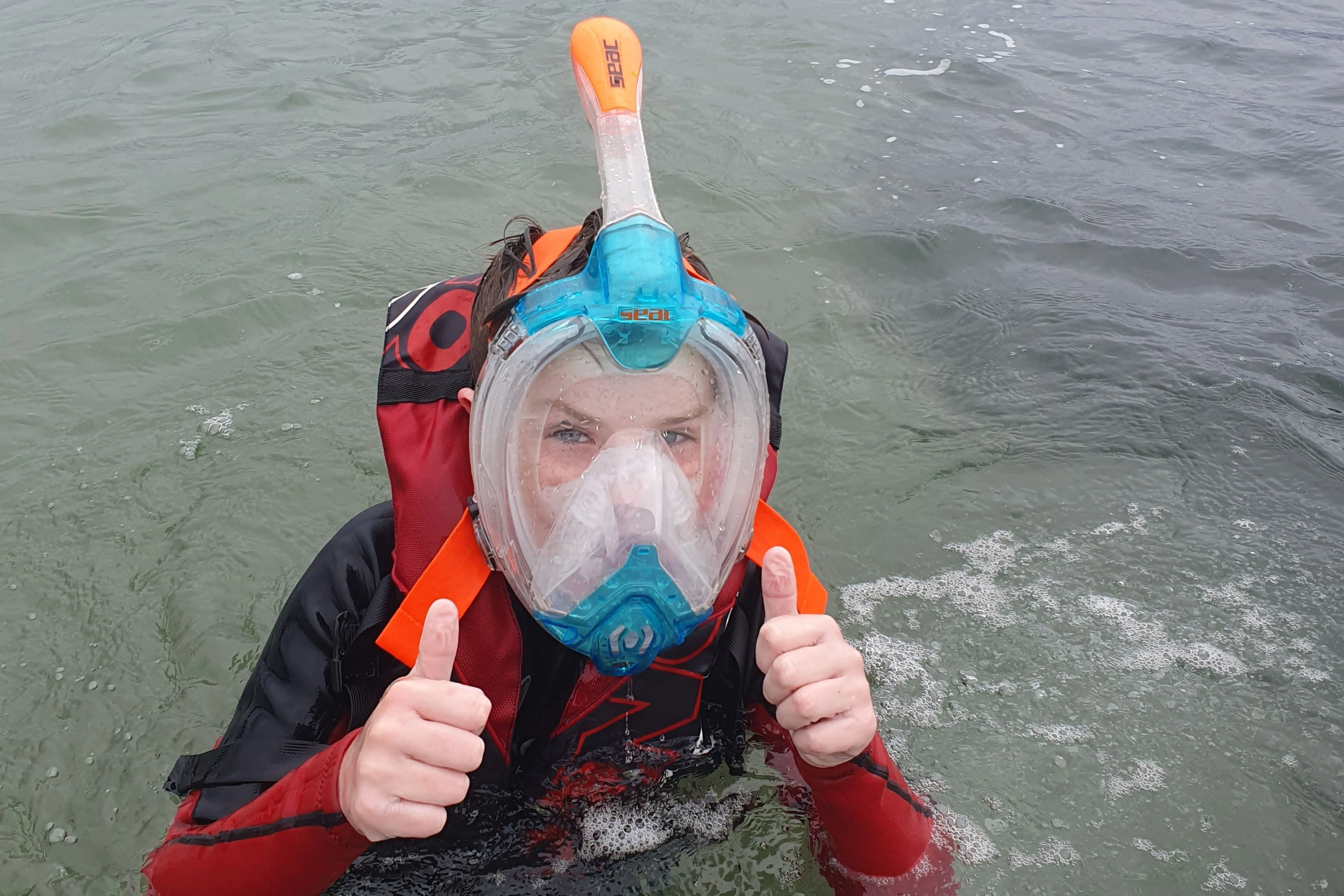 Person in water wearing a full-face snorkel mask and wetsuit giving two thumbs up.