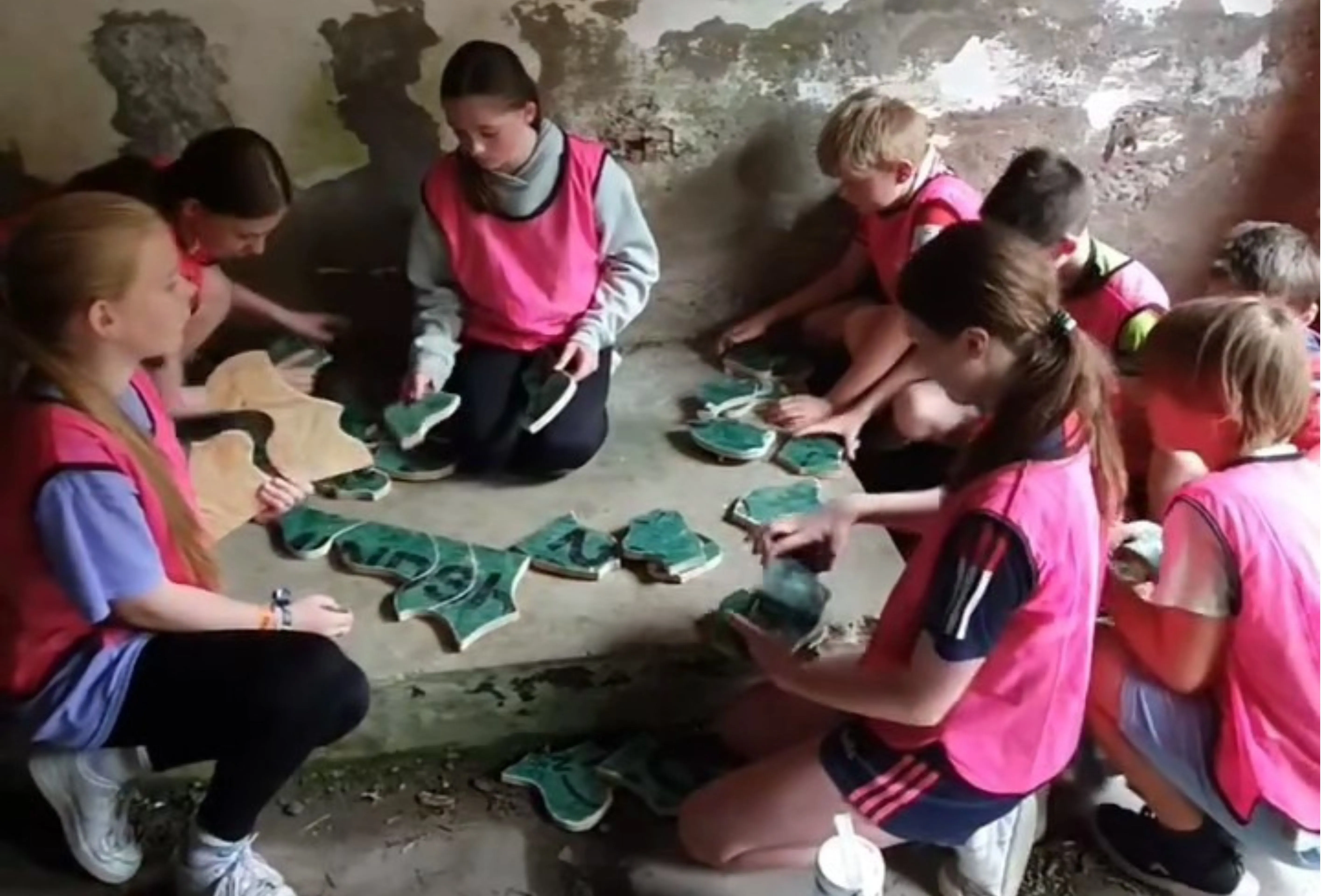Group of children wearing pink vests assembling large green puzzle pieces on a concrete surface in a dim room with worn walls.