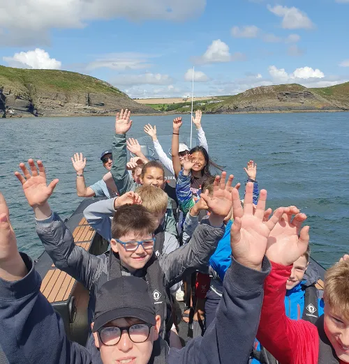 Group of smiling children on a boat raising their hands with water and green hills in the background.