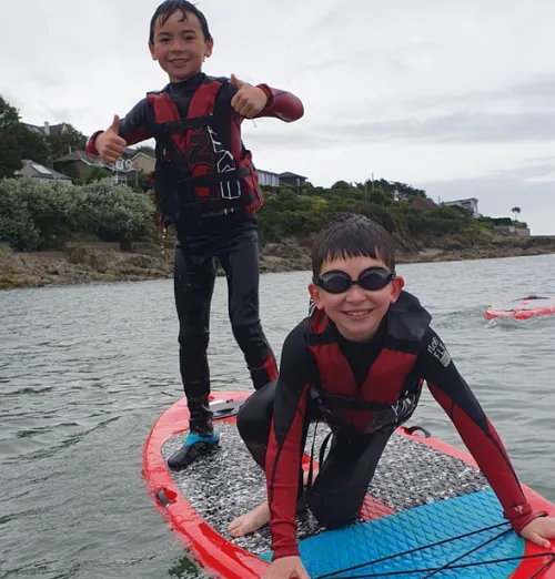 Two boys in wetsuits on a paddleboard in a calm body of water near a rocky shore with houses and greenery.