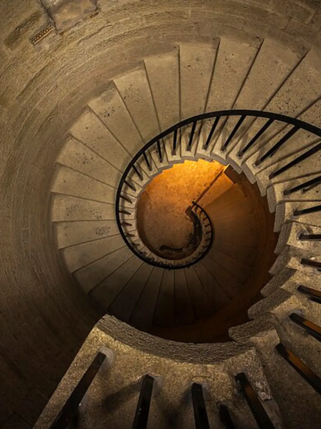 Spiral stone staircase with black metal railing viewed from the top, lit by warm light below.