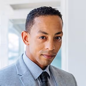 Portrait of a confident man in a light gray suit, blue shirt, and black tie against a blurred office background.