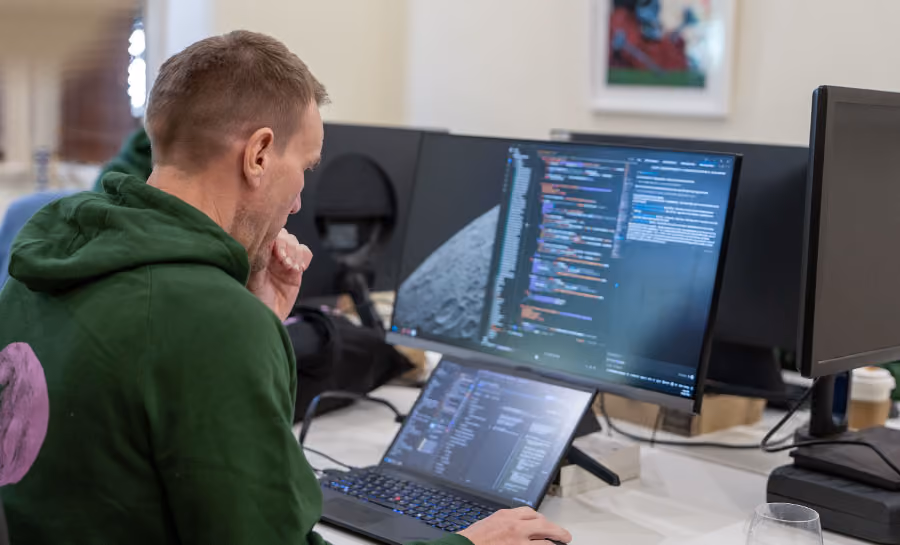 Man in green hoodie working on laptop with dual monitors displaying code and moon surface image.