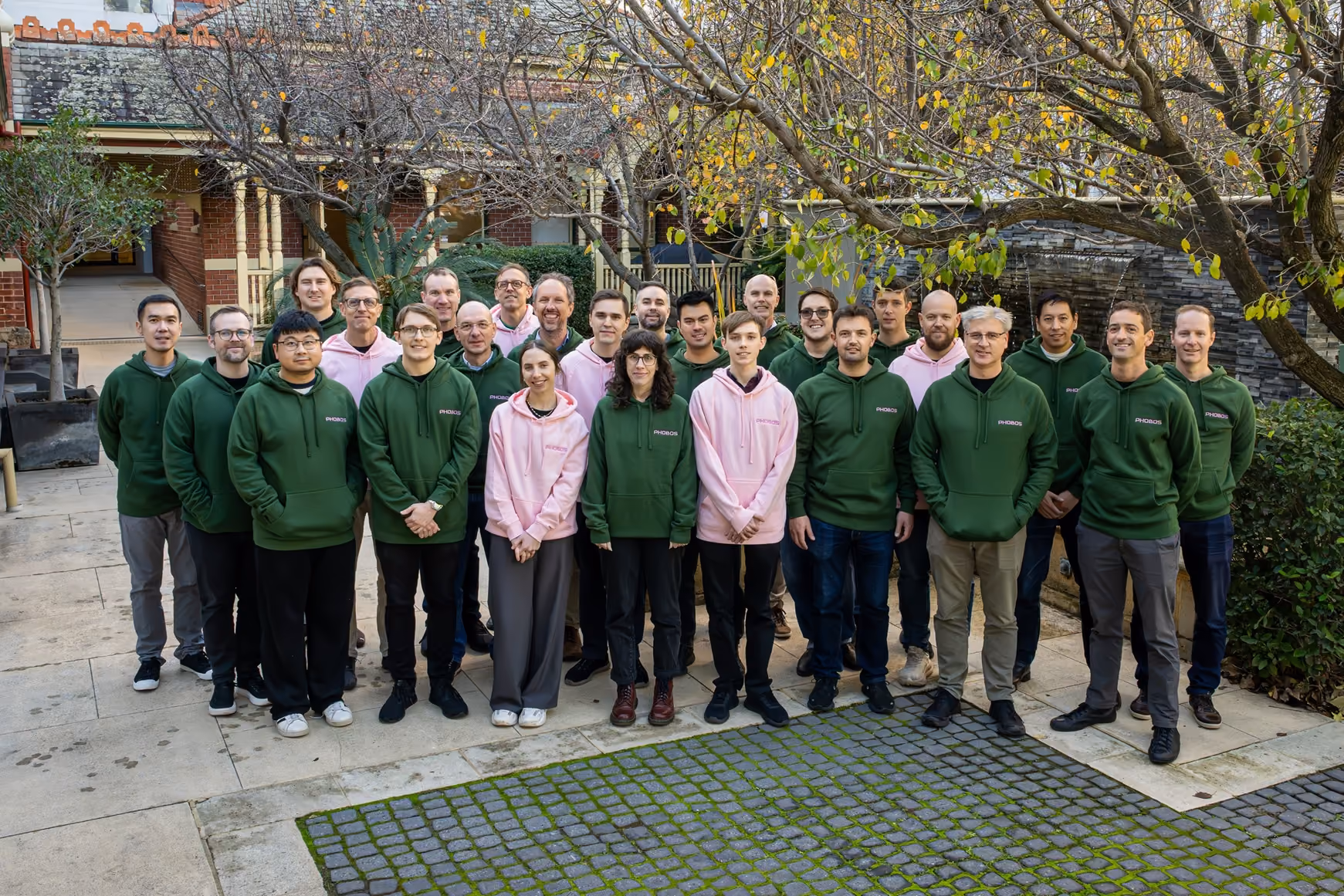 Group of 27 people standing outdoors wearing green and pink hoodies with PHOBOS logo under trees and near brick building.